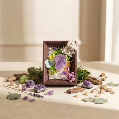 Framed amethyst landscape standing among loose crystals, moss, driftwood and stones on a sunlit table