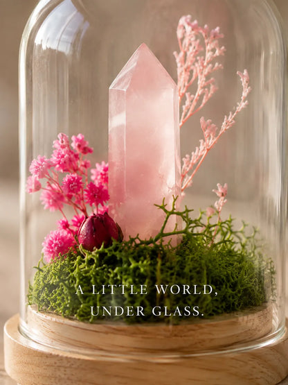 Close-up of a rose quartz point in moss and pink flowers under glass, with the line “A little world, under glass”
